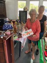 Photo of one of our vendors holding a Florida tag and a customer standing at her vendor table displaying miscellaneous household items and collectables for sale.